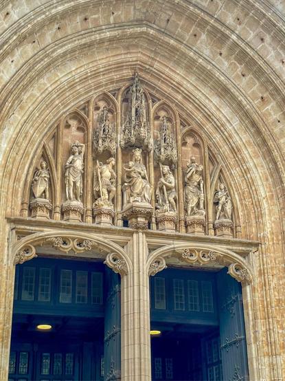 A close-up view of a grand, intricately detailed Gothic church entrance, featuring a large archway with sculptures. The facade is adorned with stone carvings, and the stairway leading up to the doors is visible.
