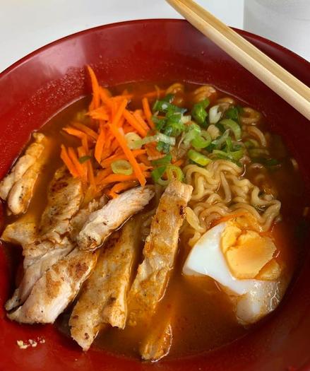 A close-up shot of a red bowl filled with ramen noodles in broth, chicken slices, shredded carrots, green onions, and half a boiled egg, with chopsticks resting on the rim of the bowl.