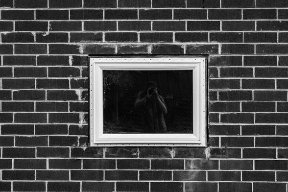 A black and white photo of a window in a brick wall with a reflection of me in the glass holding the camera to my face.