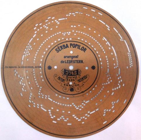 photo of a museum object, a kind of round player piano disc with holes stamped in it in a chain working its way around the disc in several layers. the label in the middle says Serba Popilor, Arangeat de Leop. Stern.