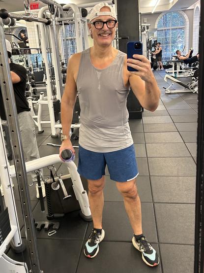 A smiling person (me)  in a gray tank top and blue shorts poses for a selfie in a gym, holding a water bottle. The background features gym equipment and other people working out.