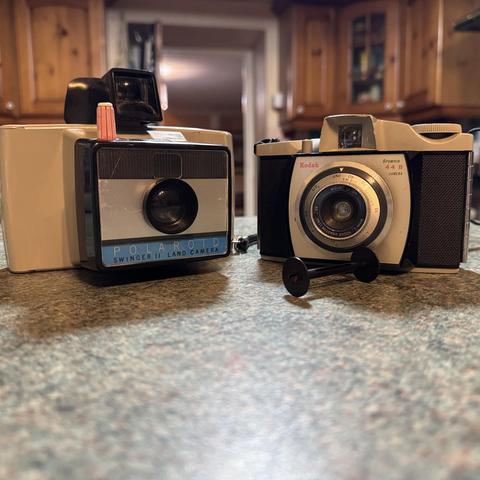 Two vintage cameras are on a kitchen surface. The camera on the right is a bit boxy affair from Polaroid called the Swinger.  The Camera on the right is a much dinkier Kodak Brownie 44B.
