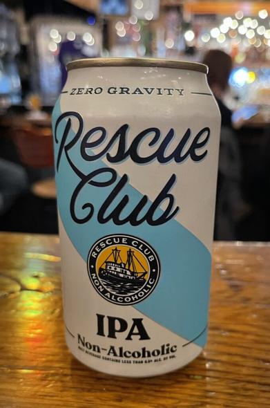 A close-up of a beer can siting on a wooden surface. It's white with a diagonal blue stripe and dark cursive lettering spelling out Rescue Club. In the background, out of focus are some people sitting at a bar. Light reflecting off of bottles etc. creates a sparkling effect.