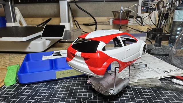 A close-up of a partially painted model car, showcasing a sleek design with a white and red color scheme, displayed on a workbench surrounded by modeling tools and materials.