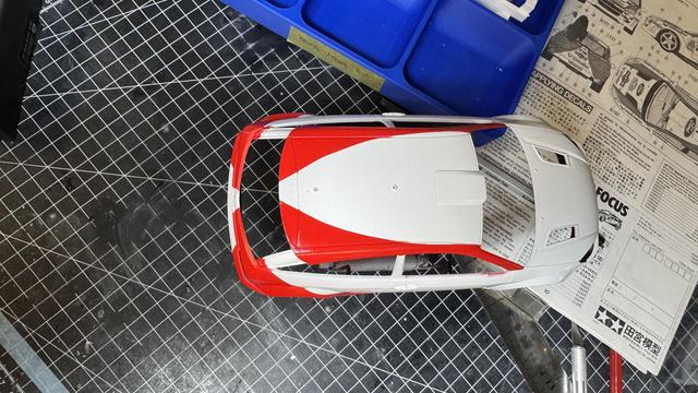 An overhead view of a model car in progress, highlighting its white and red paint job, with a blue tray of tools in the background and detailed instructions visible on the work surface.