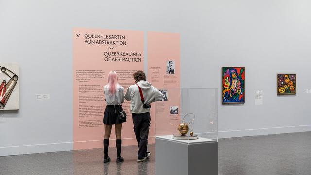 Blick in die Ausstellung. Zwei Personen stehen vor einer rosa Wand mit Text zur Ausstellung. Links und rechts davon Kunstgegenstände