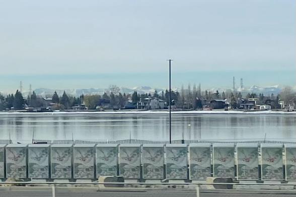 Shot from a moving car, looking across a small lake on the outskirts of Calgary towards houses on the far shore, with a dusting of snow. Way in the distance, the Rockies.