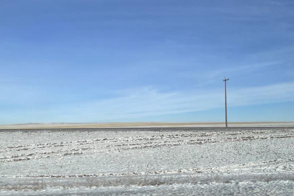 The Prairies. Mostly flat, blue sky with a few wispy clouds. In the foreground, the ground is covered with dirty snow up to a line a few hundred meters away marked with a hydro pole. Beyond that to the horizon is green.