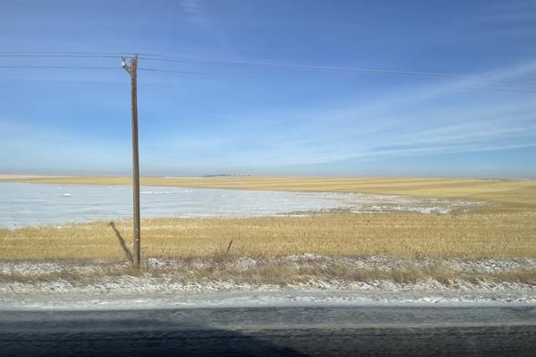 The Prairies. Mostly flat. The sky is blue with a few wispy clouds. In the foreground a telephone pole on the left of the frame. Farmland to the horizon, some covered in snow, some with what looks like the remains of crops that have been harvested.