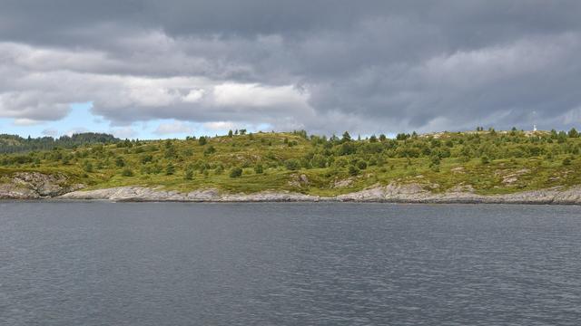 A photo of a rocky shoreline leading up a low green hill. The sky is filled with grey clouds with a little bit of blue sky exposed.