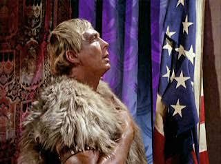 A prehistoric looking man with his hand over his heart looking at US flag