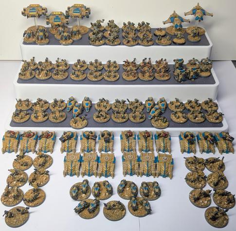 Ein Foto zeigt eine Sammlung bemalter Miniaturen eines Sci-Fi-Tabletop-Spiels, ordentlich auf mehreren weißen Stufen präsentiert. Die Figuren sind in Wüstenfarben mit blauen Akzenten bemalt und zeigen verschiedene Einheiten – Infanterie, Fahrzeuge und Fluggeräte – auf sandfarbenen Bases.