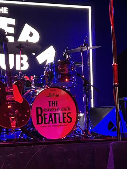 A drum set on stage with a red bass drum labeled “The Beatles,” illuminated by colorful stage lights, ready for a performance at the Cavern Club.