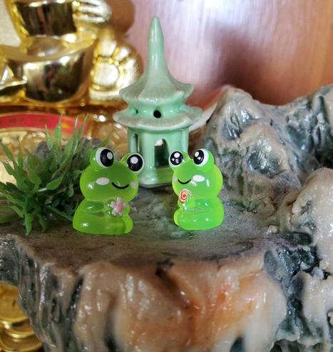 A close-up shot of an artistic miniature display. Two green, glossy frog figures are situated on an artificial rock formation, alongside a small green pagoda structure. The frog on the left holds a pink flower, while the frog on the right holds a lollipop. The setting features small artificial greenery. In the background, there is a golden statue and a backdrop.