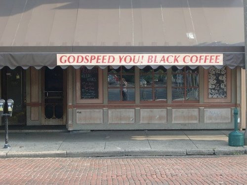 A storefront with an awning sign that says Godspeed You! Black Coffee
A play on the band named Godspeed You! Black Emperor.
There is a brick paved street in front of the store.