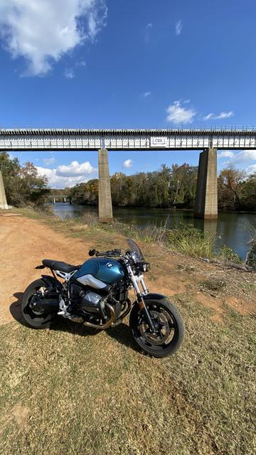 The aquamarine BMW motorcycle on the banks of the river.  Behind it is a bright blue sky and rail bridge spanning over the river.