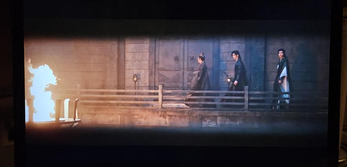 Su Mu Yu and Chang He following a man on a walkway in a stonehall. There's a brazier with a fire burning in the foreground.