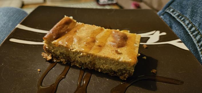 A rectangular slice of baked pumpkin cheesecake on a small, square plate, with caramel sauce drizzled across it.