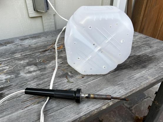A plastic milk jug has been placed on its side on an old wooden table with the bottom facing towards the camera. The bottom shows a series of small holes burned into the bottom of the jug. In the foreground is an old soldering iron.