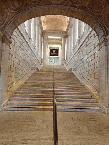 A long wide marble staircase leads up to an enormous alcove with a small pair of doors. A transom above the pair of doors shows a large chandelier in the room behind.