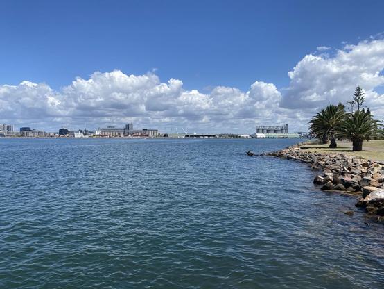 Hunter River/Newcastle Harbour seen from the Stockton side. View of some of the port/industrial facilities (such as silo-like structures) across the river from mid to right. On the left of those, newer multi-story residential buildings and several cranes no doubt working on building more apartments. Blue sky with low white clouds. Blue-grey water.