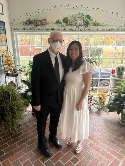 A man in a black suit wearing an N95 mask. A young woman in a white dress stands beside him.