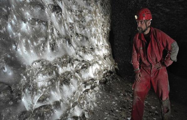 A researcher in caving gear stands near a giant colonial spiderweb on the wall of a cavern.