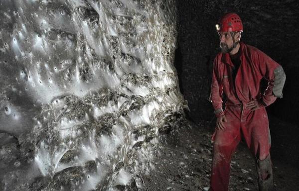 A researcher in caving gear stands near a giant colonial spiderweb on the wall of a cavern.