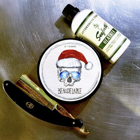 (Le Chat Mistral description)
This image shows a trio of shaving items arranged on a metallic surface.
At the top, there is a white bottle of "Seaforth! Heather Aftershave Tonic", featuring a green label with gold text.
In the centre, there is a round tin of shaving soap named "Beaudelaire", with a label depicting a skull wearing sunglasses and a red Santa hat.
At the bottom, there is a straight razor with a black handle and gold detailing, branded "H. Boker Tree Brand", partially unfolded to reveal its metallic blade.