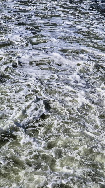 Es handelt sich um die Aufnahme einer ruhelosen und schäumenden Wasseroberfläche mit weißer Gischt. Es vermischen sich Grüntöne mit dunklen und hellen 
Farbtönen.