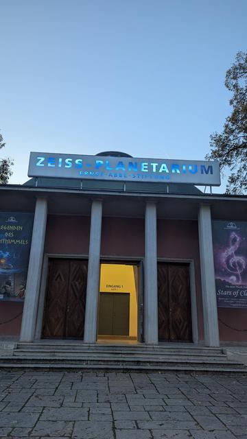 Blick aufs Zeiss Planetarium in Jena von außen. Rechts und links hängen banner zu Veranstaltungen und oben der Schriftzug vom Planetarium in Jena.