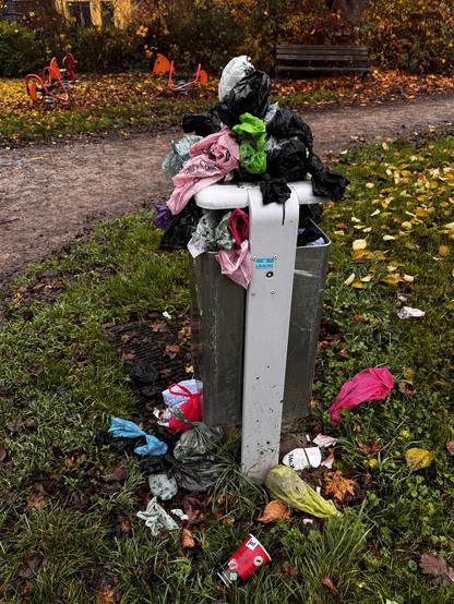 Ein überquellender Mülleimer, gefüllt mit verschiedenen Hundekotbeuteln, darunter schwarze, rosa, grüne und blaue. Einige Abfälle liegen verstreut um den Mülleimer herum, und im Hintergrund sind Spielgeräte und abgefallene Blätter zu sehen.