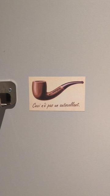 Un piccolo sticker attaccato a una parete mostra il disegno della famosa pipa di Magritte e la scritta in francese “Ceci n’est pas un autocollant.”, cioè “Questo non è un adesivo.”