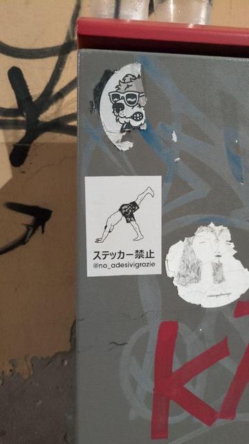 Un’immagine mostra un armadietto o una cabina metallica grigia, con alcuni adesivi attaccati sopra. Tra questi, al centro, c’è un adesivo bianco con il disegno stilizzato di una persona in posizione capovolta, come se stesse facendo una verticale o una piroetta. Sotto al disegno ci sono delle scritte in giapponese (“ステッカー禁止”, che significa “vietato attaccare adesivi”) e l’handle di Instagram “@no_adesivigrazie”. Sullo sfondo si vedono altri adesivi parzialmente strappati e dei graffiti neri sul muro.