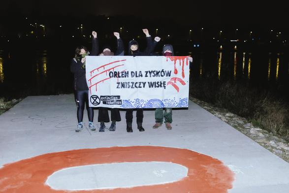 4 aktywistki XR nocą na tle Wisły, trzymają baner z napisem "Orlen dla zysków zniszczy Wisłę". Przed nimi na betonowym podłożu namalowana kilkumetrowa litera "O".