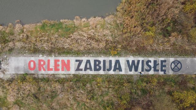 Zdjęcie z powietrza ostrogi na Wiśle, porośniętej krzewami i drzewami. Pośrodku droga wyłożona betonowymi płytami, na której namalowano kilkumetrowej wysokości napis "Orlen zabija Wisłę".