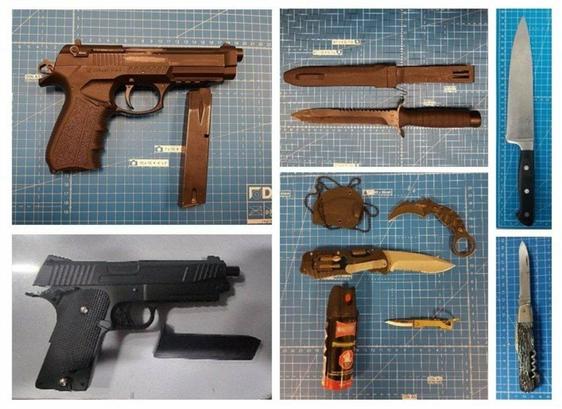 Das Bild zeigt eine Sammlung von Waffen, die auf einer blau karierten Schneidematte liegen. Zu sehen sind zwei Pistolen, diverse Messer, ein Pfefferspray und ein kleines Holster. Eine der Pistolen ist eine schwarze Pistole mit herausgenommenem Magazin. Die zweite Pistole ist ebenfalls schwarz und hat ein Magazin, das daneben liegt. Bei den Messern handelt es sich um verschiedene Typen, darunter ein großes Küchenmesser, ein feststehendes Messer mit gezackter Klinge, ein Klappmesser und ein Karambit-Messer. Das Pfefferspray ist rot und schwarz.