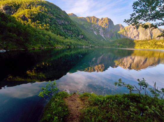 The image depicts a serene landscape featuring a calm lake surrounded by lush green mountains. The water is remarkably still, creating a perfect mirror image of the surrounding scenery, including the mountains and the sky. The mountains are steep and rugged, with patches of greenery and exposed rock, indicating a natural, untouched environment. The vegetation on the mountains is dense, with trees and shrubs covering the slopes. The sky is partly cloudy, with soft, diffused light suggesting either early morning or late afternoon. In the foreground, there is a small patch of grass and a few young plants, with a narrow path leading towards the water's edge. The overall color palette is dominated by greens, blues, and earthy tones, contributing to the tranquil atmosphere of the scene.