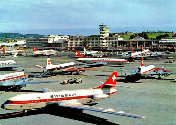 A vibrant postcard view of Zürich Airport (Kloten) from the late 1950s or early 1960s shows a bustling ramp filled with gleaming Swissair aircraft in their classic red-and-white livery. The scene captures the optimism of the early jet age — polished metal fuselages reflecting sunlight under a crisp alpine sky.

In the foreground, a Sud Aviation Caravelle, with its distinctive rear-mounted engines and swept wings, prepares for departure. Its fuselage bears the bold SWISSAIR name and a red stripe of porthole windows accented by the white cross of Switzerland on the tail. Behind it, several Douglas DC-6 and Convair aircraft sit at gates, their propellers still, as ground crews and luggage carts move about.

The modernist terminal building rises in the background, a long rectangular structure with horizontal bands of windows and a control tower centered above. Beyond the airport, green hills and scattered trees signal the Swiss countryside.

This image evokes a golden era of European aviation — a time when Swissair proudly called itself “The Airline of Switzerland,” and Zürich stood as one of the Continent’s most elegant and efficient gateways to the world.