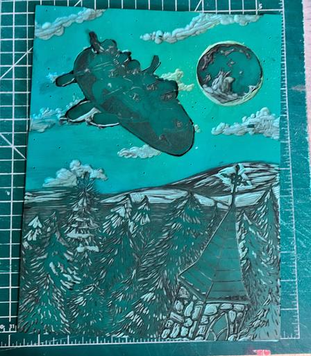 A carved green vinyl Lino block on a green cutting mat shows a night sky with moon and clouds and flying canoe full of paddling voyageurs over rolling countryside with fields and trees covered in snow and a church steeple in the foreground