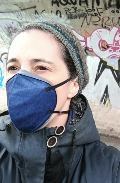a white woman stands in front of a wall full of graffiti and tags. She is wearing a grey and blue toque, a dark blue coat and a blue respirator.