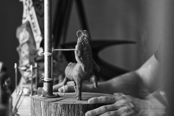 Black and white photograph of a clay sculpture on a wooden podium. The sculpture depicts a centaur with horns, in an upright posture.