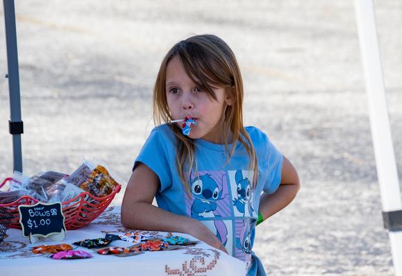 A young girl with a Lilo & Stitch shirt on and her elbow resting on a vendor's table.  She has a lollypop in her mouth with the wrapper still on the stem, apparently so she doesn't have to finish it and can rewrap it for later.