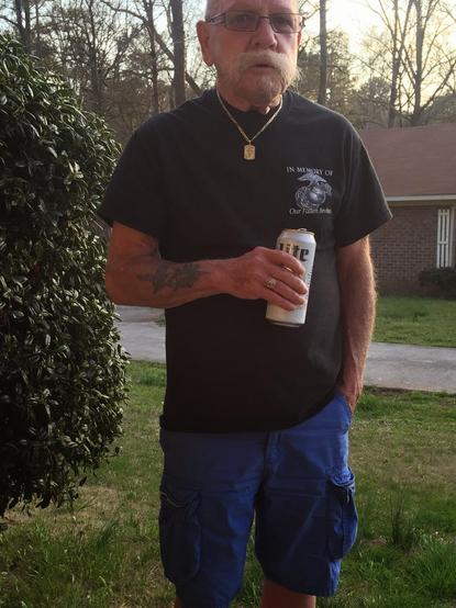 A photo of my dad, Pops, holding a Miller Lite beer can, black Marine Corps t-shirt, blue shorts with a gold necklace around his neck. You can see part of a house in the background as well as a bush and some grass.