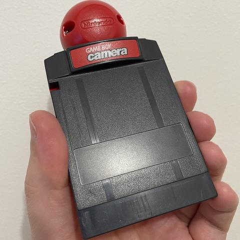 A person holds a Game Boy Camera, a small accessory for capturing photos. It features a red lens on top and a prominent label reading "GAME BOY camera." The device has a compact, rectangular shape with a smooth black surface.