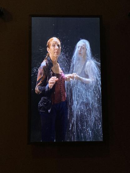Two people walking through a waterfall in a black studio, holding hands. The first one has already passed and is pictured in colours, the second one is just stepping through water, seen in grayish tones.