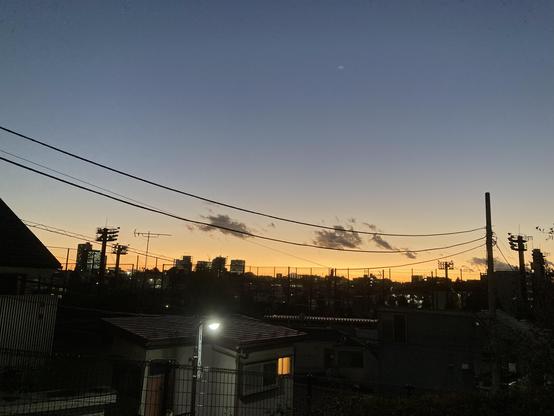 A view of evening city. Silhouettes of black houses huddling against the background of clear, wintery-looking sky. Orangey (slightly golden)  gloaming can be seen near the horizon and above is an empty, darker-blue space.
