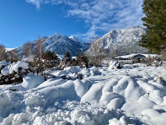 The landscape after a snowstorm has soft mounds of snow accumulated in front of houses, almost to the point that the houses are not visible. There are a few scattered trees, and beyond the houses and trees is the Sierra Nevada mountain range, covered in snow. The sky is blue with some spotty clouds. Sheridan, Nevada in the Carson Valley