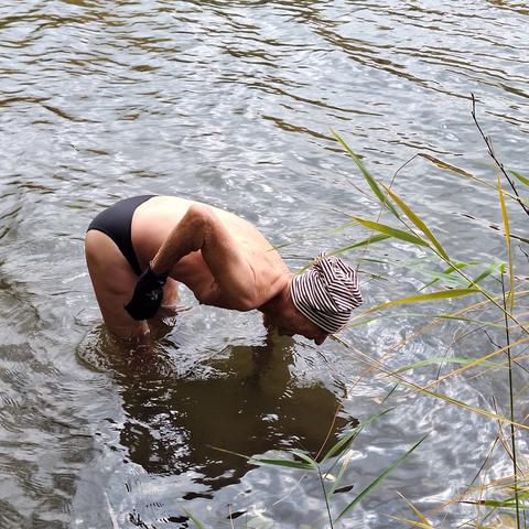 Ein Mann in schwarzer Badehose und braun-beige gestreifter Mütze steht bis zu den Knien im Wasser und streckt den linken Arm hinein. Der rechte Arm wird nach oben angewinkelt rausgehalten. An der rechten Hand trägt er einen Neopren-Handschuh.
Am rechten Bildrand sind vereinzelte Schilfrohrhalme und ein dünner Zweig zu sehen.
Die Wasseroberfläche ist leicht gewellt, blassblau, hell- und dunkelgrau im Wechsel von hell und dunkel.