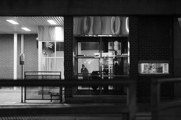 The station exterior at night, looking south. Far left are the lights of the outdoor platform, but no riders are visible. Centre is the large window revealing the glass windows of the elevator and beyond into the station, one person moving through the station. At far right is one small, horizontal window revealing a glimpse of more passengers.